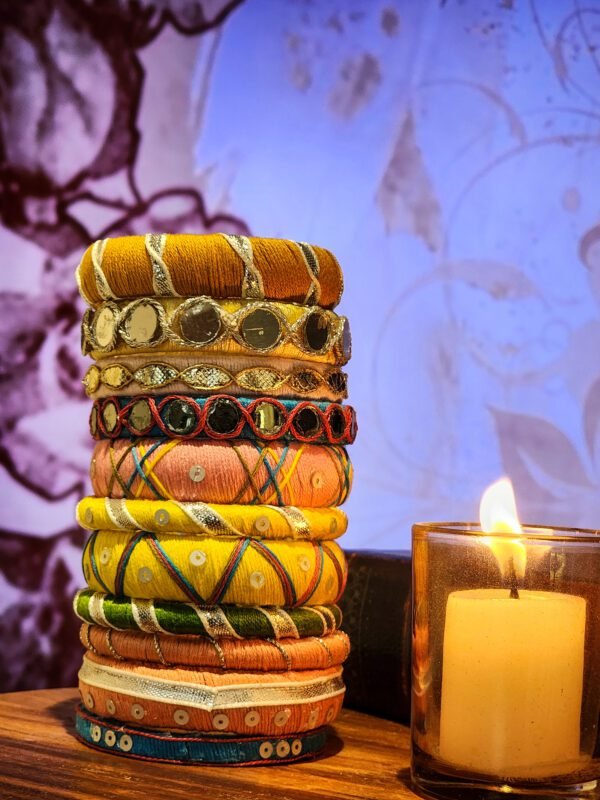 A stack of handcrafted thread bangles in gold, red, blue, and green, adorned with mirrorwork, sequins, and intricate embroidery, displayed on a wooden surface beside a flickering candle. The warm glow highlights the artisanal craftsmanship of South Asian textiles.