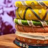 A stack of handcrafted thread bangles in gold, red, blue, and green, adorned with mirrorwork, sequins, and intricate embroidery, displayed on a wooden surface beside a flickering candle. The warm glow highlights the artisanal craftsmanship of South Asian textiles.