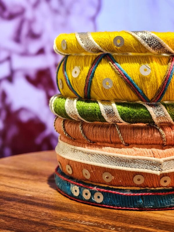 A stack of handcrafted thread bangles in gold, red, blue, and green, adorned with mirrorwork, sequins, and intricate embroidery, displayed on a wooden surface beside a flickering candle. The warm glow highlights the artisanal craftsmanship of South Asian textiles.