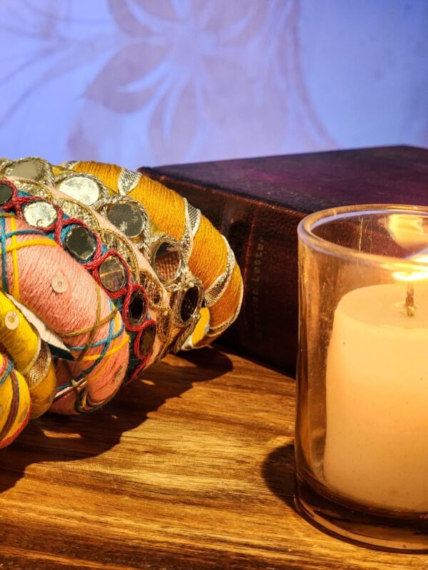 A stack of handcrafted thread bangles in gold, red, blue, and green, adorned with mirrorwork, sequins, and intricate embroidery, displayed on a wooden surface beside a flickering candle. The warm glow highlights the artisanal craftsmanship of South Asian textiles.