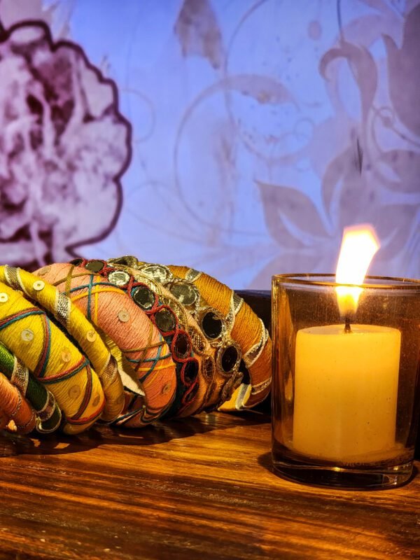 A stack of handcrafted thread bangles in gold, red, blue, and green, adorned with mirrorwork, sequins, and intricate embroidery, displayed on a wooden surface beside a flickering candle. The warm glow highlights the artisanal craftsmanship of South Asian textiles.